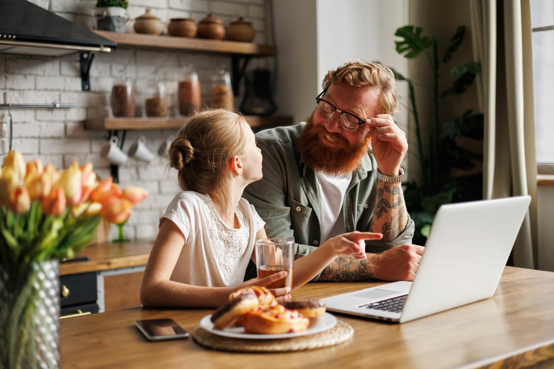 Ein Mann sitzt mit seiner Tochter am Küchentresen vor einem Laptop. Sie unterhalten sich. Die Tochter zeigt auf den Bildschirm.