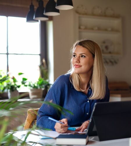 Eine blonde Frau sitzt vor ihrem Laptop, macht sich Notizen und schaut verträumt zur Seite.