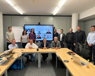 Project Labs in Linz Menschen sitzen und stehen um einen Tisch herum und lächeln in die Kamera. Im Hintergrund ist ein großer Bildschirm, auf dem weitere Menschen iun einer digitalen Konferenz zugeschaltet sind.