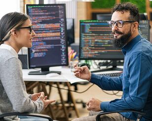 zwei Personen vor mehreren Monitoren mit Code diskutieren miteinander