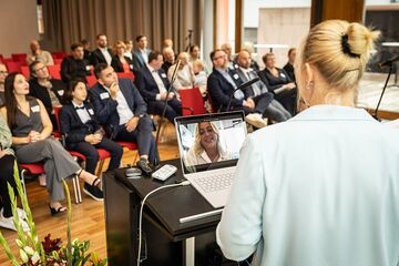 Rednerin an Pult mit Laptop und zugeschalteter Frau auf Bildschirm; im Hintergrund Publikum sitzend.