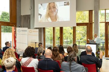 Sitzendes Publikum mit Blick auf Leinwand mit einer zugeschalteten Frau.