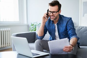 Junger Mann mit Unterlagen in der Hand, Handy am Ohr und Notebook auf Tisch lässt sich beraten.