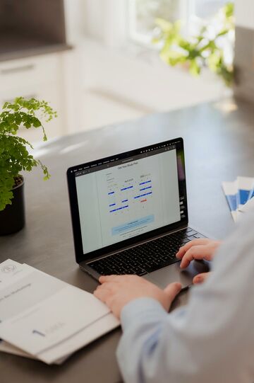 Laptop auf Schreibtisch mit Unterlagen daneben und halbsichtbarer sitzender Person