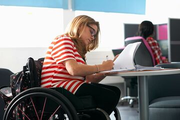 Junge Frau im Rollstuhl sitzt an Schreibtisch und schreibt in Ordner