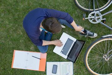 ein junger Mann lernt auf Wiese mit Laptop und gedruckten Unterlagen